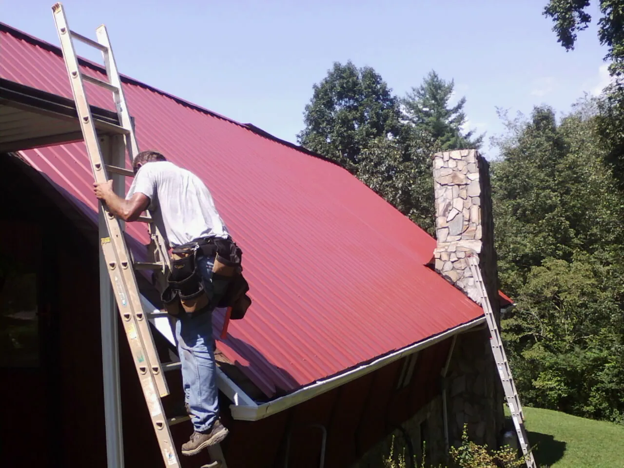 Roofer on a ladder beside a red metal roof and stone chimney — Soffit & Fascia Repair by Roofing Brothers in Milroy