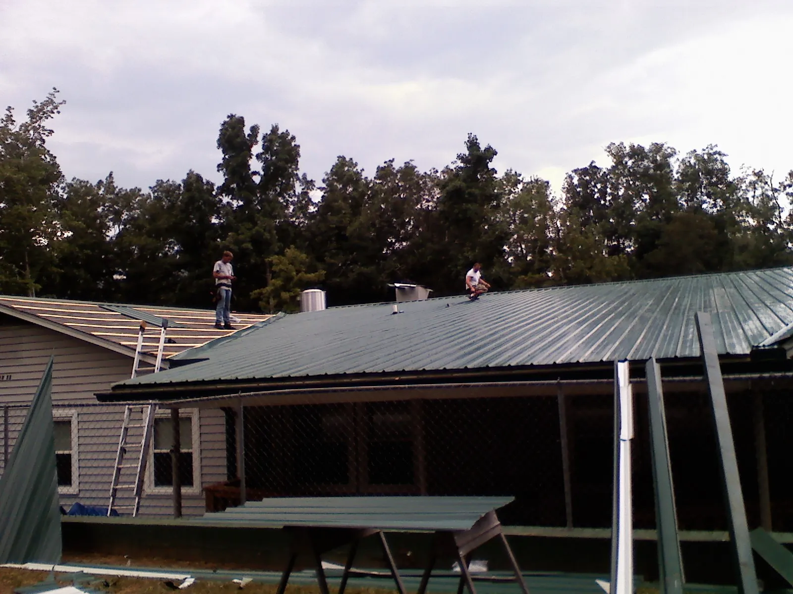 Two roofers installing green metal panels above a covered porch in Milroy